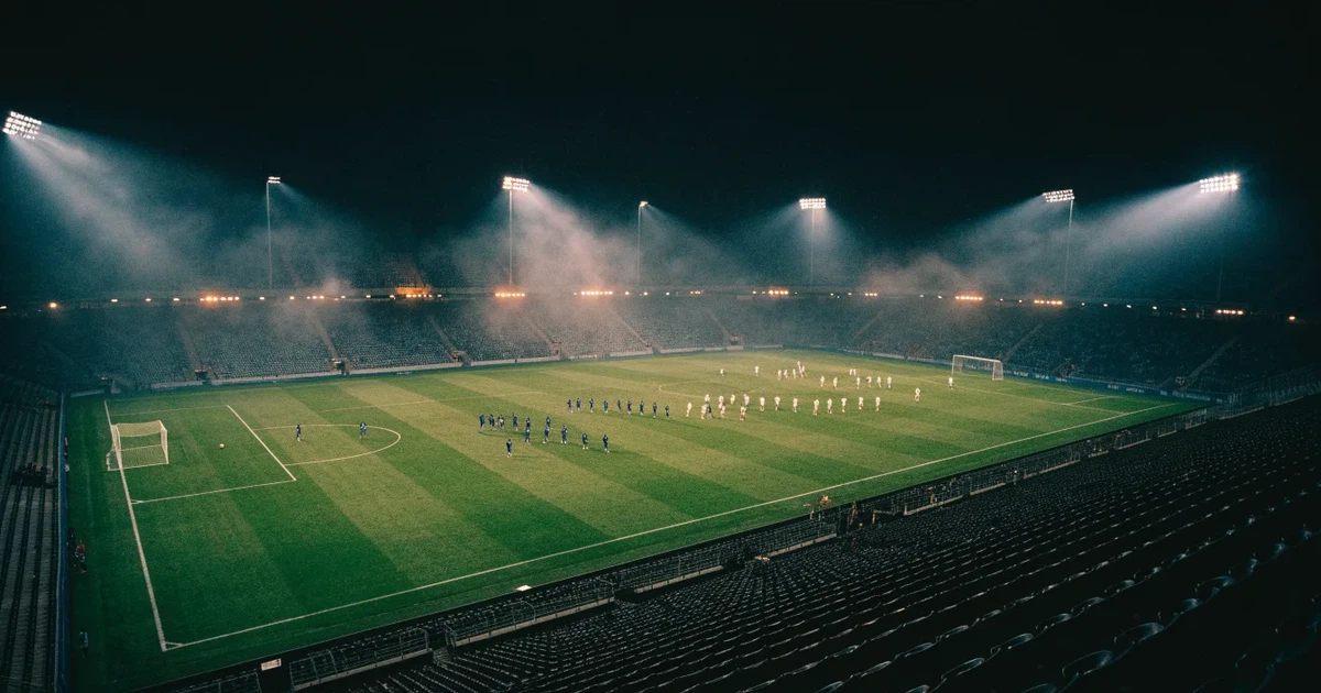 Ein Fussballspiel am Abend aus der Tribünenperspektive, die Spieler sind nur als kleine Figuren zu erkennen und das Flutlicht beleuchtet den nassen Rasen.