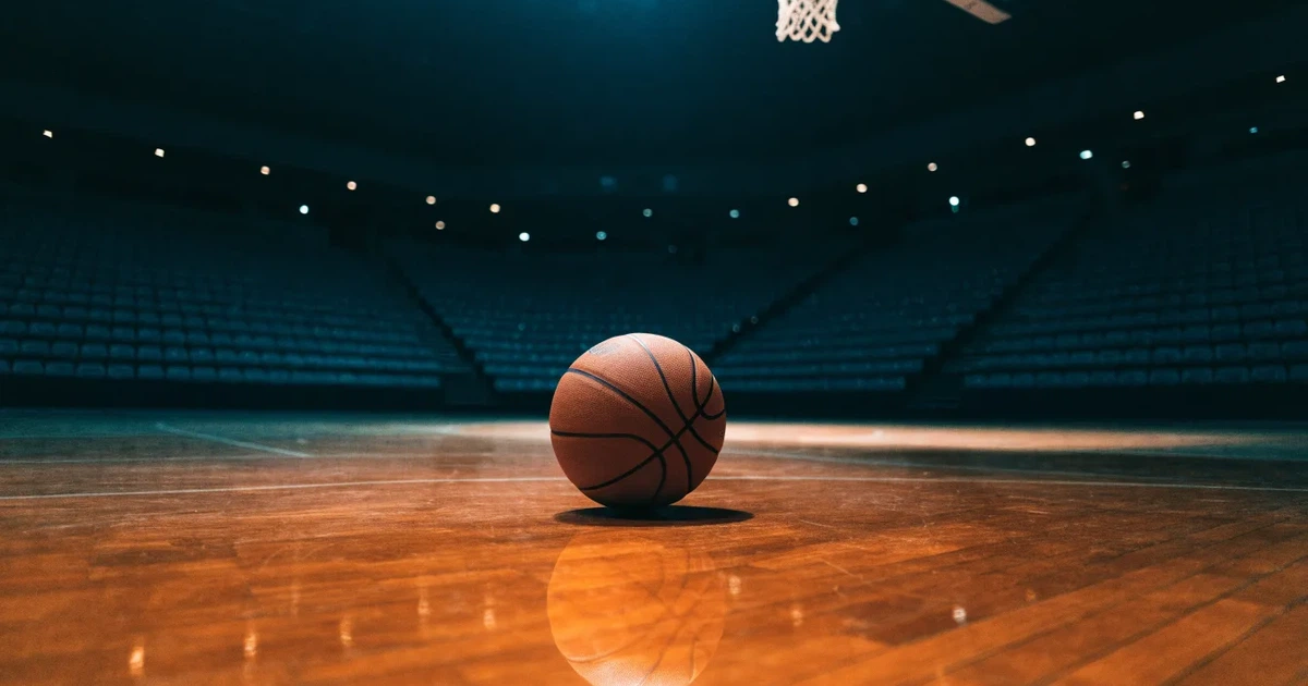 Ein Basketball liegt auf dem hellen Parkett einer leeren Halle, im Hintergrund steigen die Tribünenreihen im Halbdunkel an.
