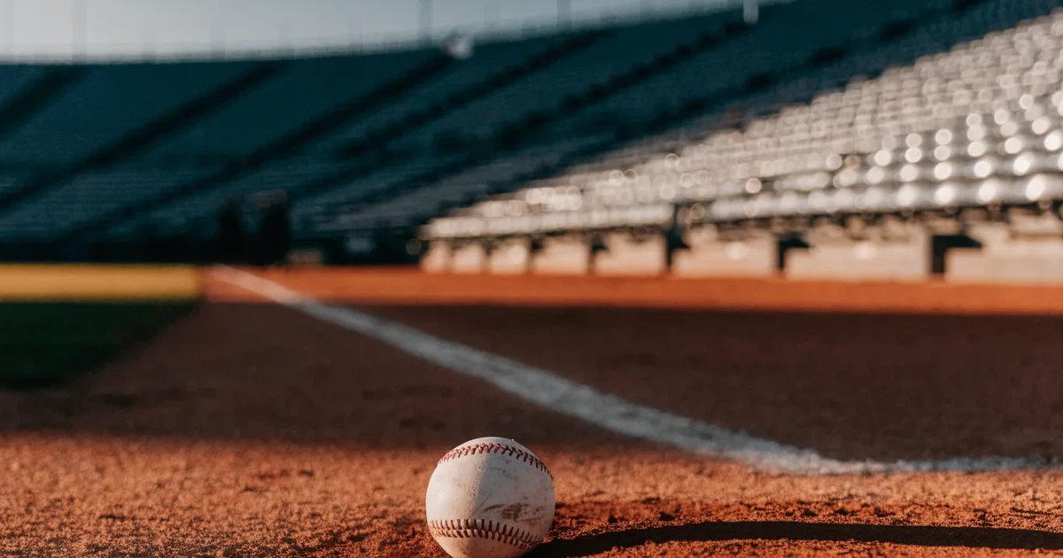 Ein weisser Baseball liegt auf rotbraunem Sand neben der Grundlinie eines leeren Stadions im warmen Abendlicht, die Ränge sind verlassen.