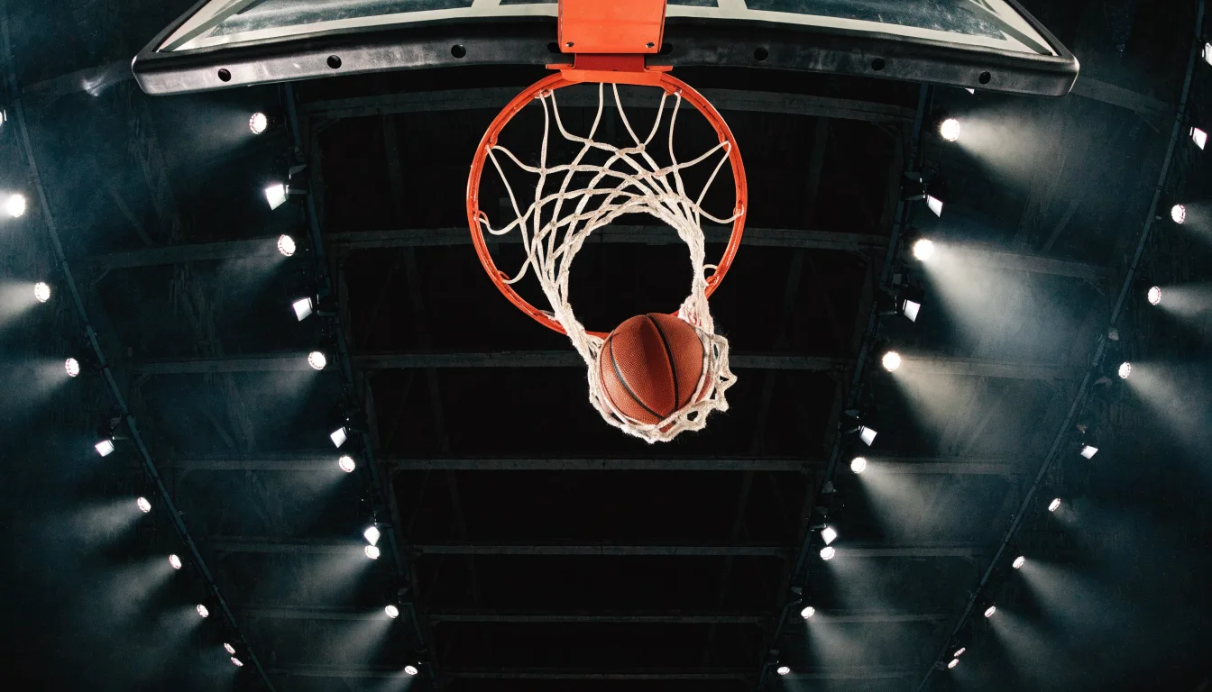 Ein Basketballkorb von unten gesehen, darüber eine Hallendecke mit Scheinwerfern und Stahlträgern.