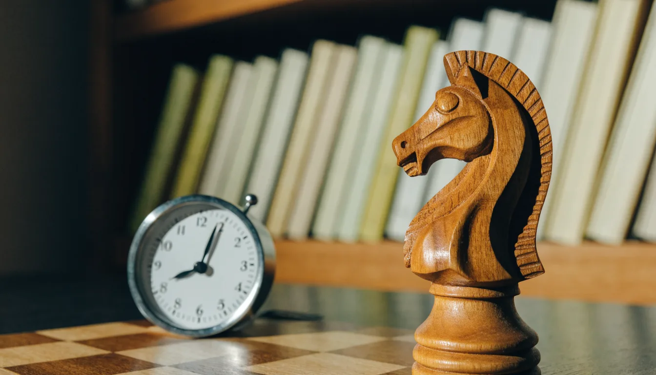 Ein Springer aus Holz steht allein auf einem Brett neben einer Schachuhr aus Metall, im Hintergrund unscharfe Bücherregale.
