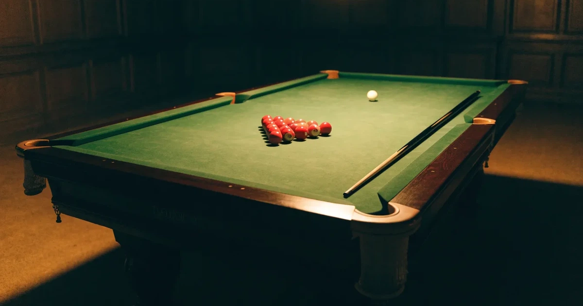 Ein Snookertisch mit grünem Tuch und einem Dreieck aus roten Kugeln unter einer tief hängenden Lampe in einer leeren Halle.