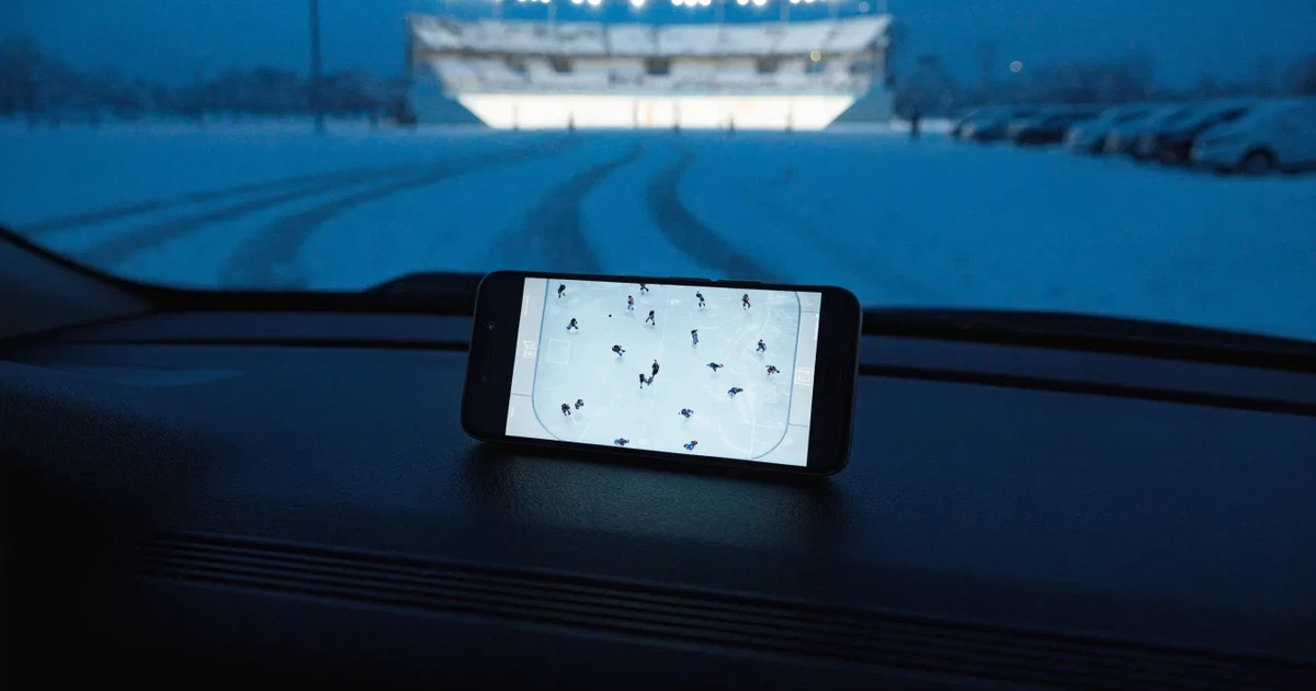 Eine Eishalle steht in der Dämmerung unter Flutlicht, aus der Ferne gesehen, davor ein leerer Parkplatz mit Schnee.