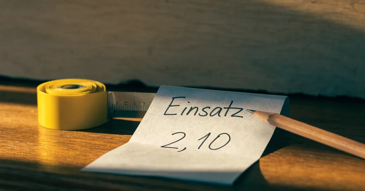 Ein Massband liegt aufgerollt auf einer Werkbank neben einem Bleistift und einem Holzbrett.